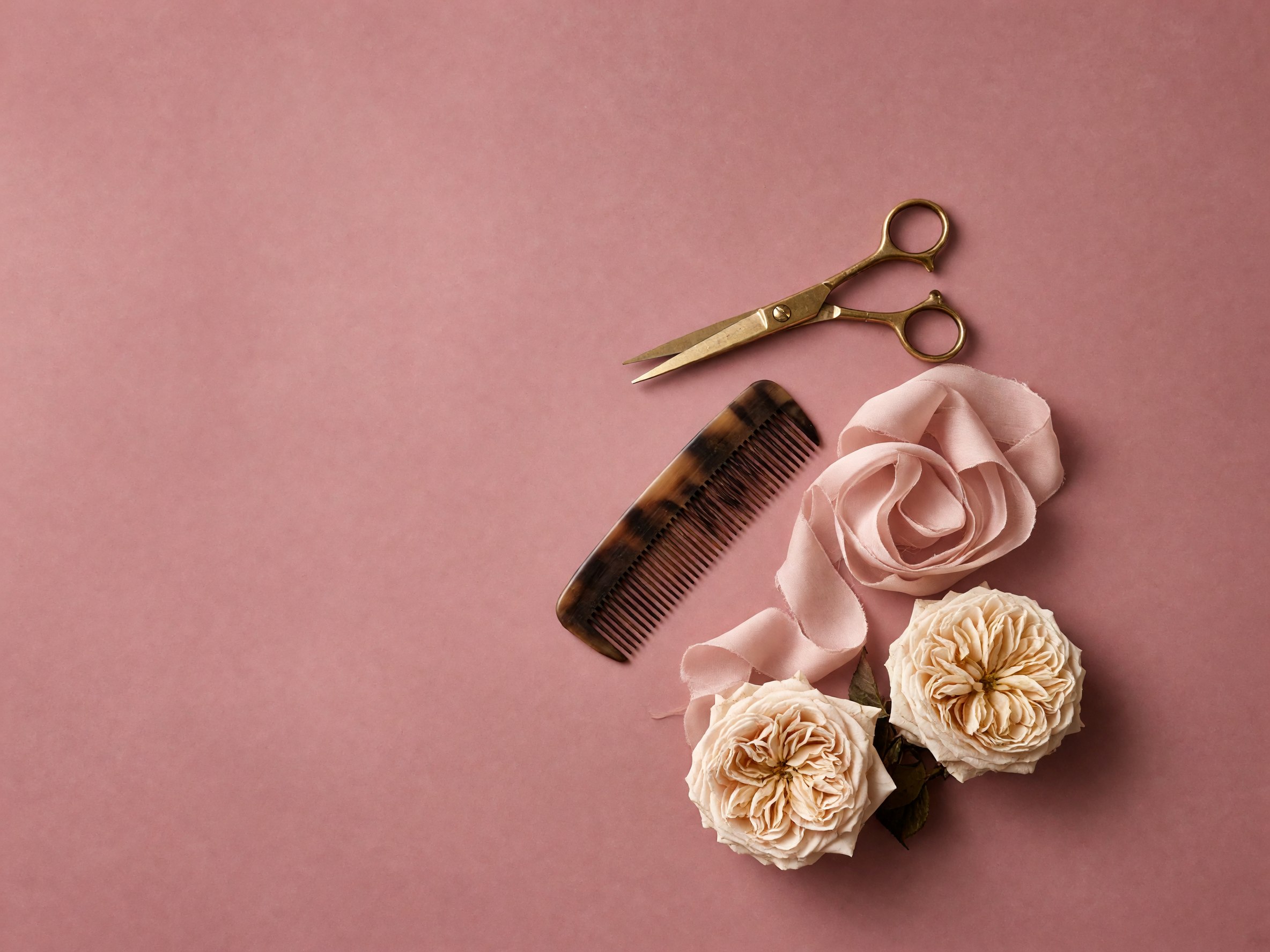 Gold scissors, a horn comb, silk ribbon, and cream roses on dusty rose paper.